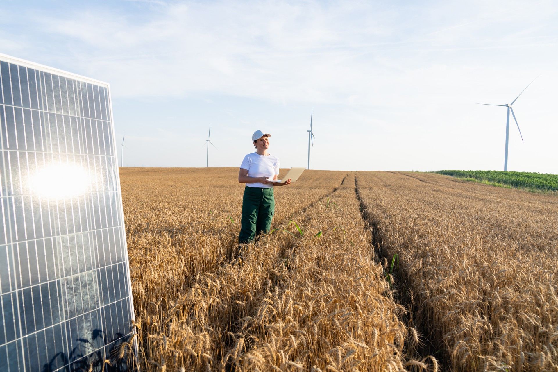 Woman farmer wearing white cap and t-shirt with laptop works next to solar panel. Wind turbines in the background